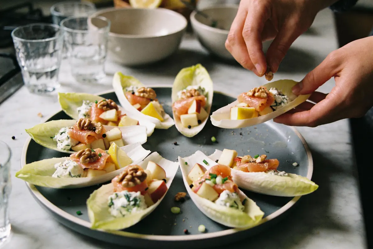 Assiette de feuilles d'endive garnies de fromage, saumon, noix et pommes, sur table avec mains ajoutant ingrédients.