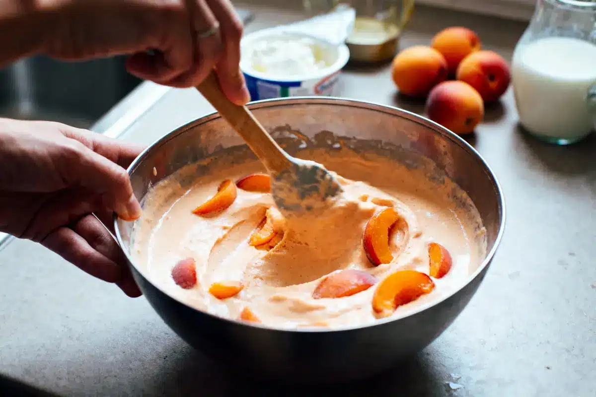 Mains mélangeant pâte avec morceaux d'abricots dans un bol en métal, sur plan de travail.