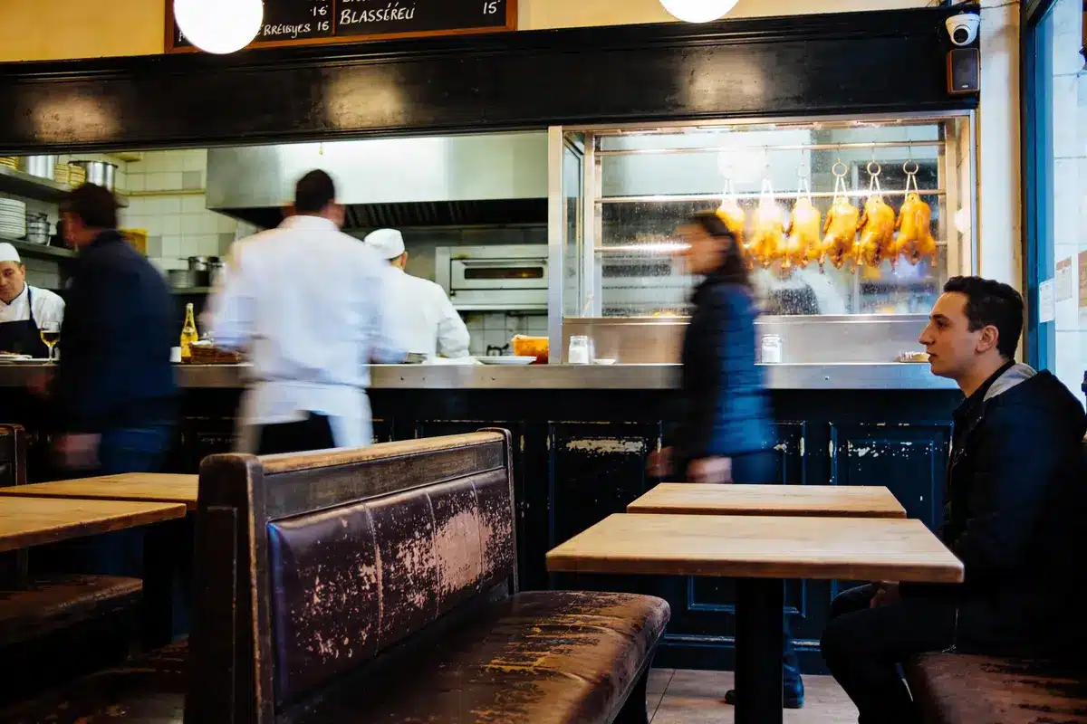 Restaurant animé avec des chefs cuisinant, personnes passant, canards rôtis suspendus, et homme assis sur une banquette.