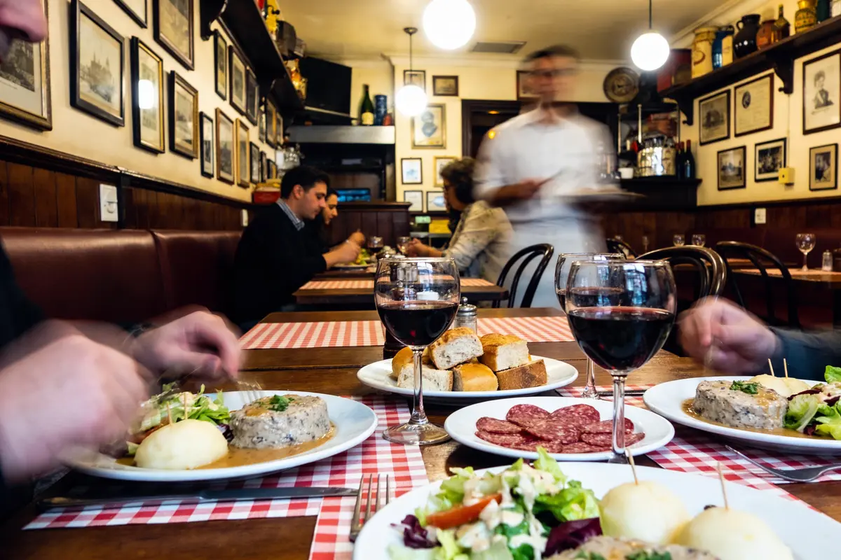 Assiettes de nourriture, verres de vin, et serveur flou dans un restaurant animé à l'intérieur chaleureux.