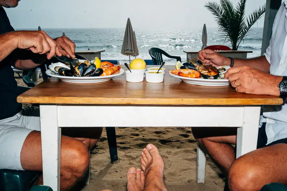Deux personnes dégustent des fruits de mer sur la plage, pied visible en premier plan, mer en arrière-plan.