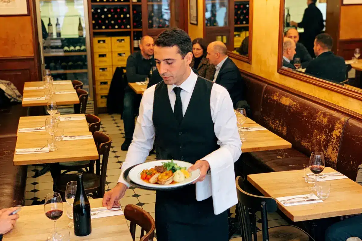 Serveur en uniforme portant plat dans un restaurant élégamment décoré avec clients en arrière-plan.