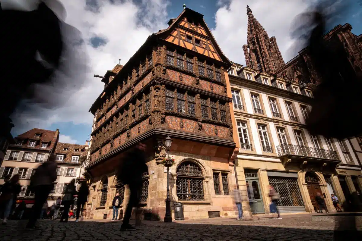 Maison médiévale ornée, typique de Strasbourg, avec passants flous, sous un ciel partiellement nuageux.