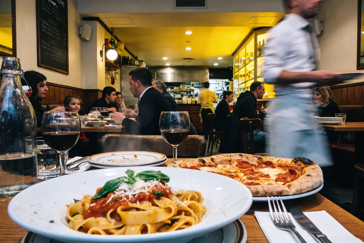 Restaurant animé, pizzas et pâtes sur table, clients discutant, serveur en mouvement, ambiance conviviale, lumière chaude.