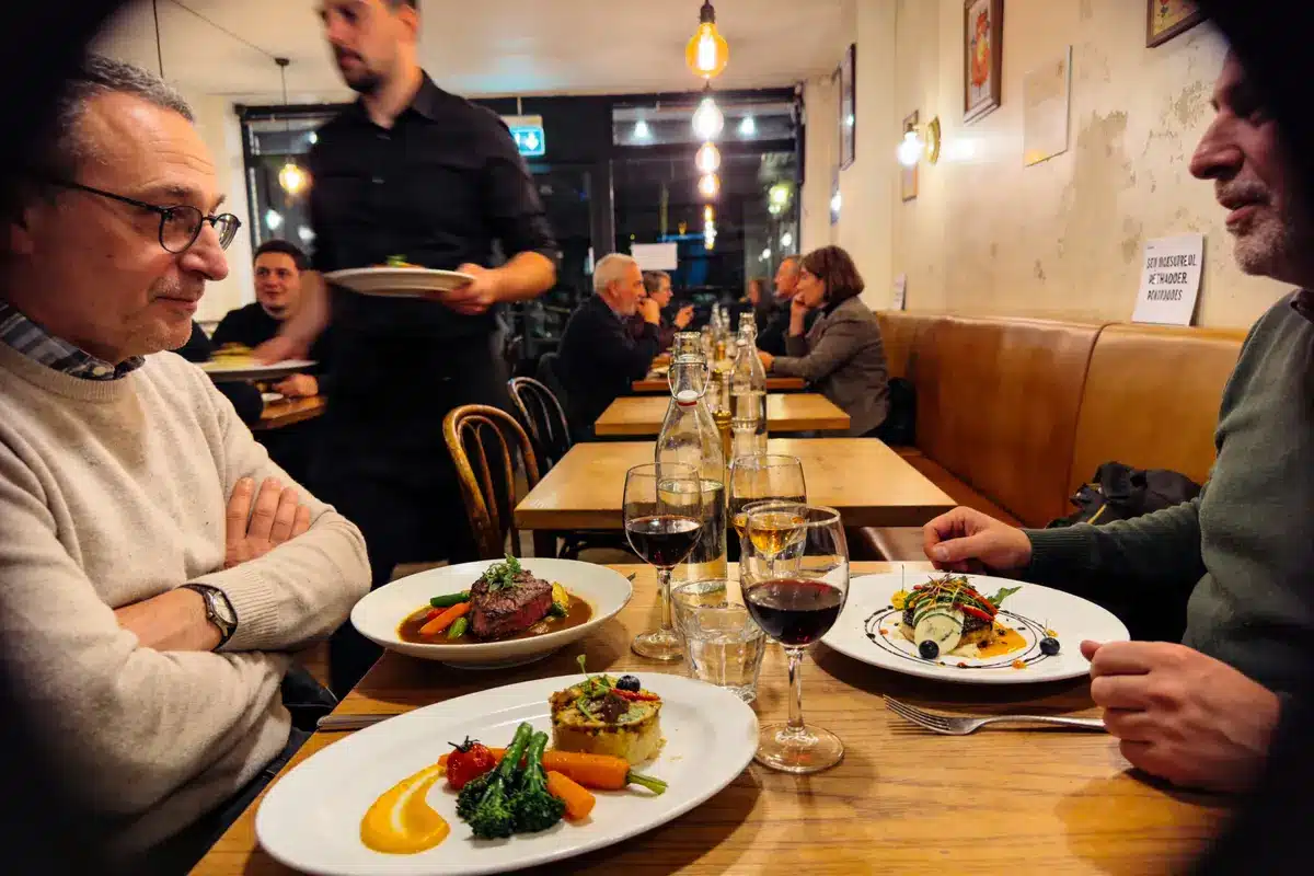 Deux hommes dans un restaurant calme, dégustant des plats élégants, avec du vin. Serveur en arrière-plan.