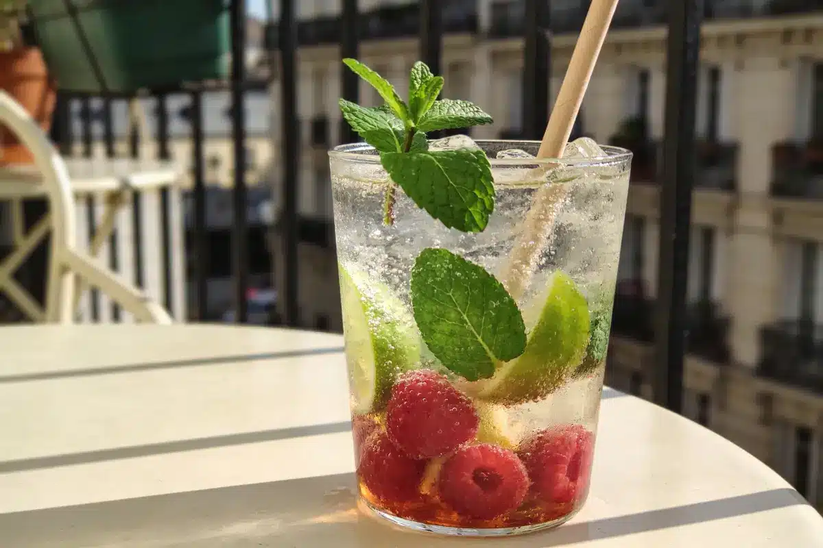 Verre de cocktail avec framboises, citron vert, menthe, et paille sur une table en extérieur.