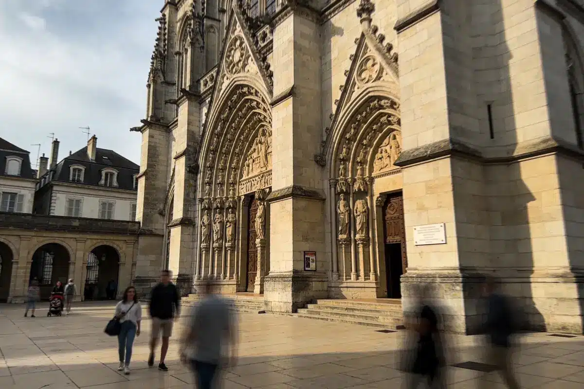 Cathédrale gothique avec sculptures et portes en bois, passants flous au premier plan, bâtiments historiques adjacents.