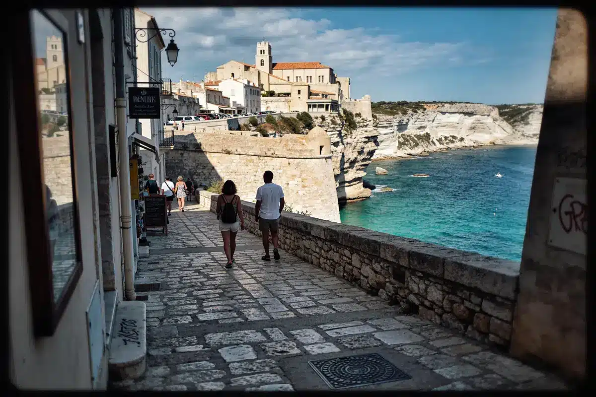 Rue piétonne pavée à Bonifacio, vue mer et falaises. Église en arrière-plan, ciel bleu.