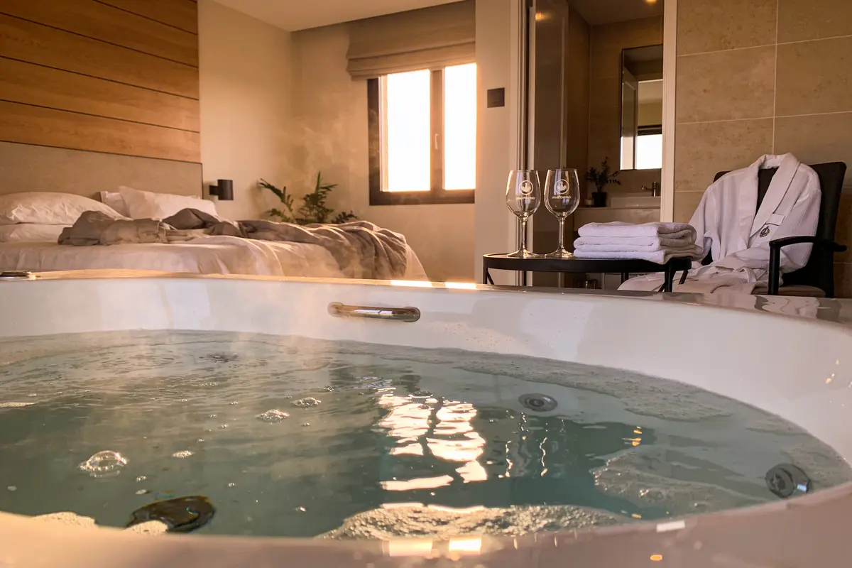 Chambre d'hôtel avec jacuzzi, lit double, fenêtre, deux verres à vin, peignoir et serviettes sur chaise.